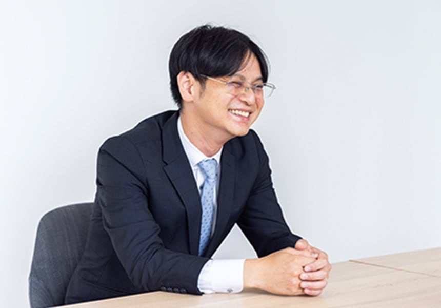 スーツ姿で笑顔の男性が会議テーブルで手を組んで座っている様子